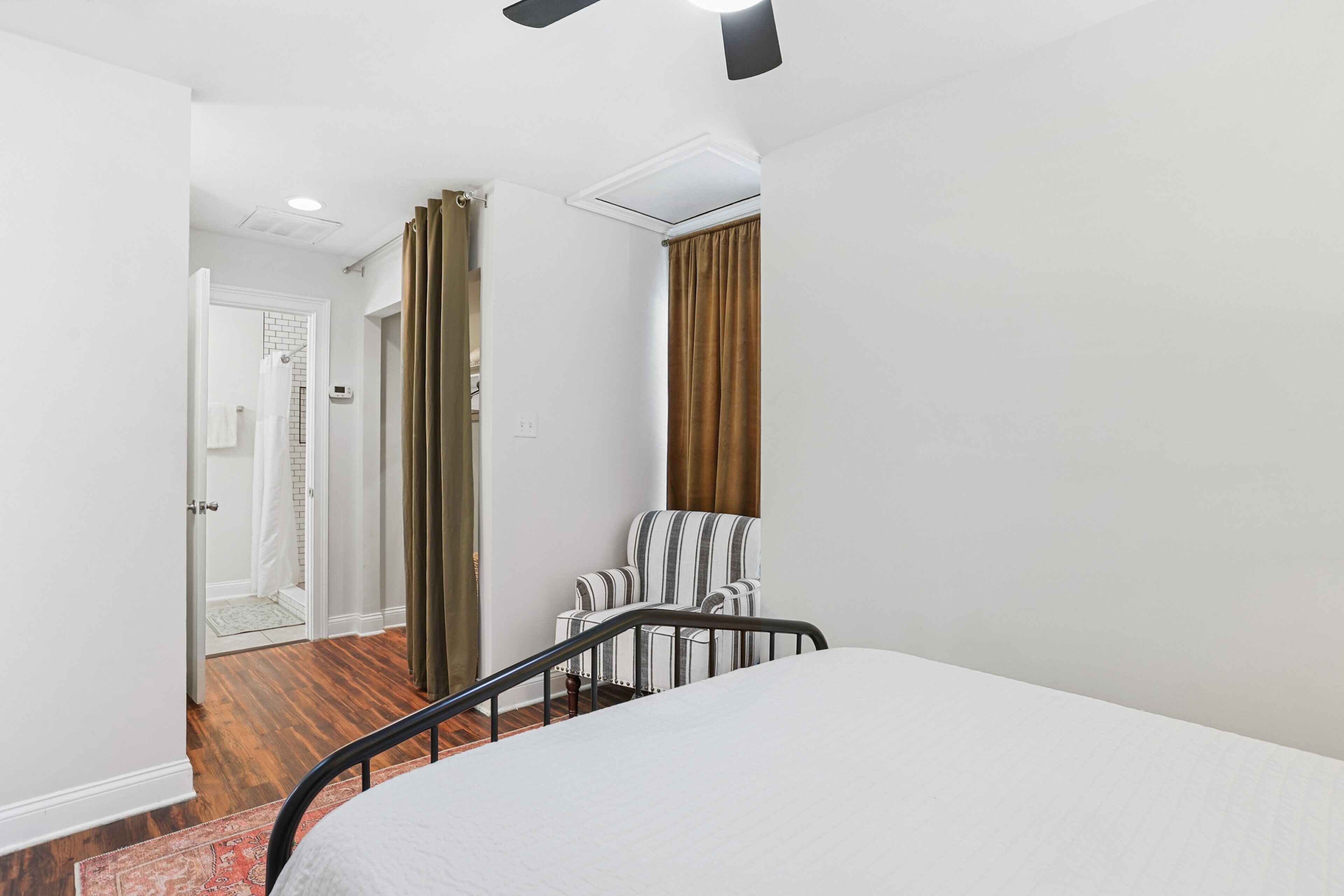 A bedroom with a bed, a chair, a window with curtains, a door, and a ceiling fan.