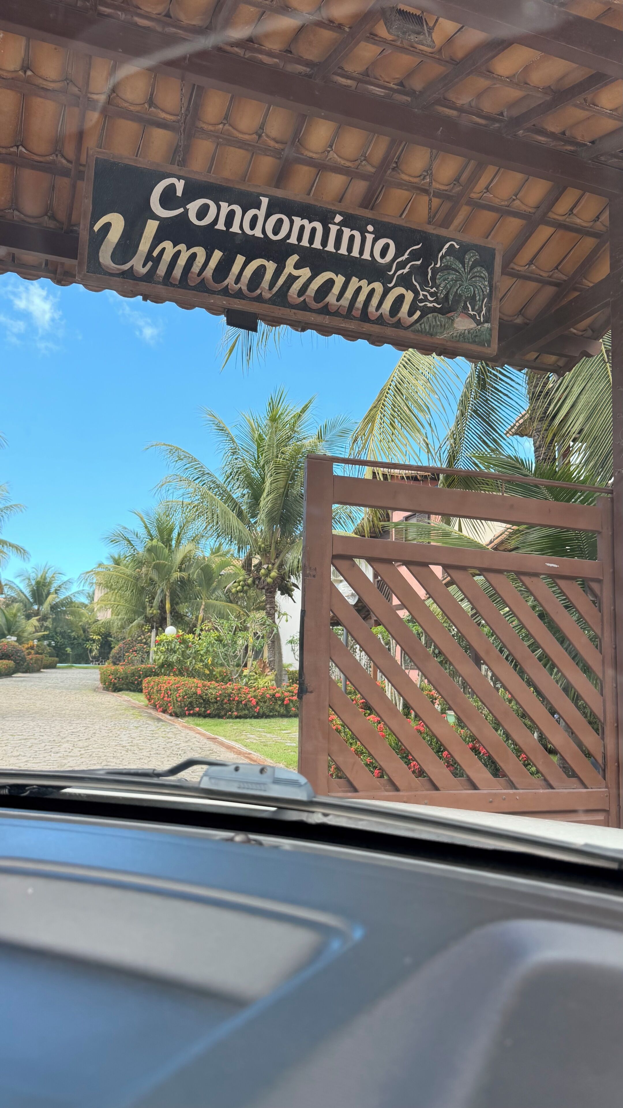 A gated entrance with a sign reading 'Condominio Umuarama' and a view of palm trees and a cobblestone path. - Premium House | 250m Taperapuan Beach | Axé Moi (Porto Seguro)