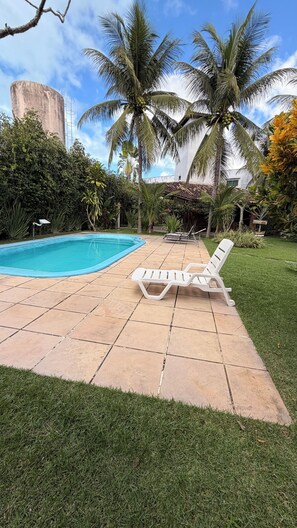 A swimming pool with a tiled deck, surrounded by palm trees and a grassy area with lounge chairs. - Premium House | 250m Taperapuan Beach | Axé Moi (Porto Seguro)