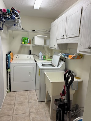 A laundry room with a washing machine, dryer, and iron. There are shelves with cleaning supplies and a vacuum cleaner. - Vacation Rental Property by S.Gondola Dr with private swimming pool (Venice)