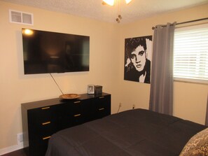 A bedroom with a bed, a dresser, a television mounted on the wall, and a window with curtains. - Three Kings Retreat - East Memphis (Memphis)