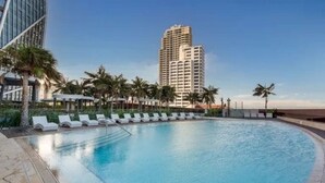 A swimming pool with lounge chairs, palm trees, and a tall building in the background. - Opulence Oceanview 2BR Suite- The Langham + Parking (Surfers Paradise)