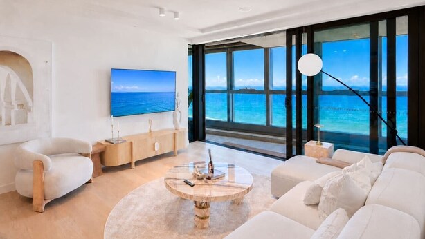 A modern living room with a large window offering a view of the ocean, a flat-screen TV mounted on the wall, and a round coffee table with decorative items. - Opulence Oceanview 2BR Suite- The Langham + Parking (Surfers Paradise)