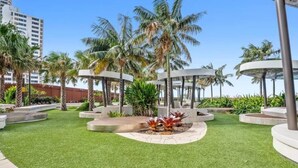 A landscaped outdoor area with palm trees, a circular seating area, and a modern building in the background. - Opulence Oceanview 2BR Suite- The Langham + Parking (Surfers Paradise)