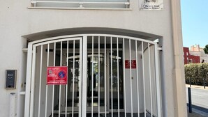 Un edificio con entrada protegida por una reja, un letrero que indica 'Port Bressou 1' y 'Est. 100', y una señal de 'Prohibido estacionar'.