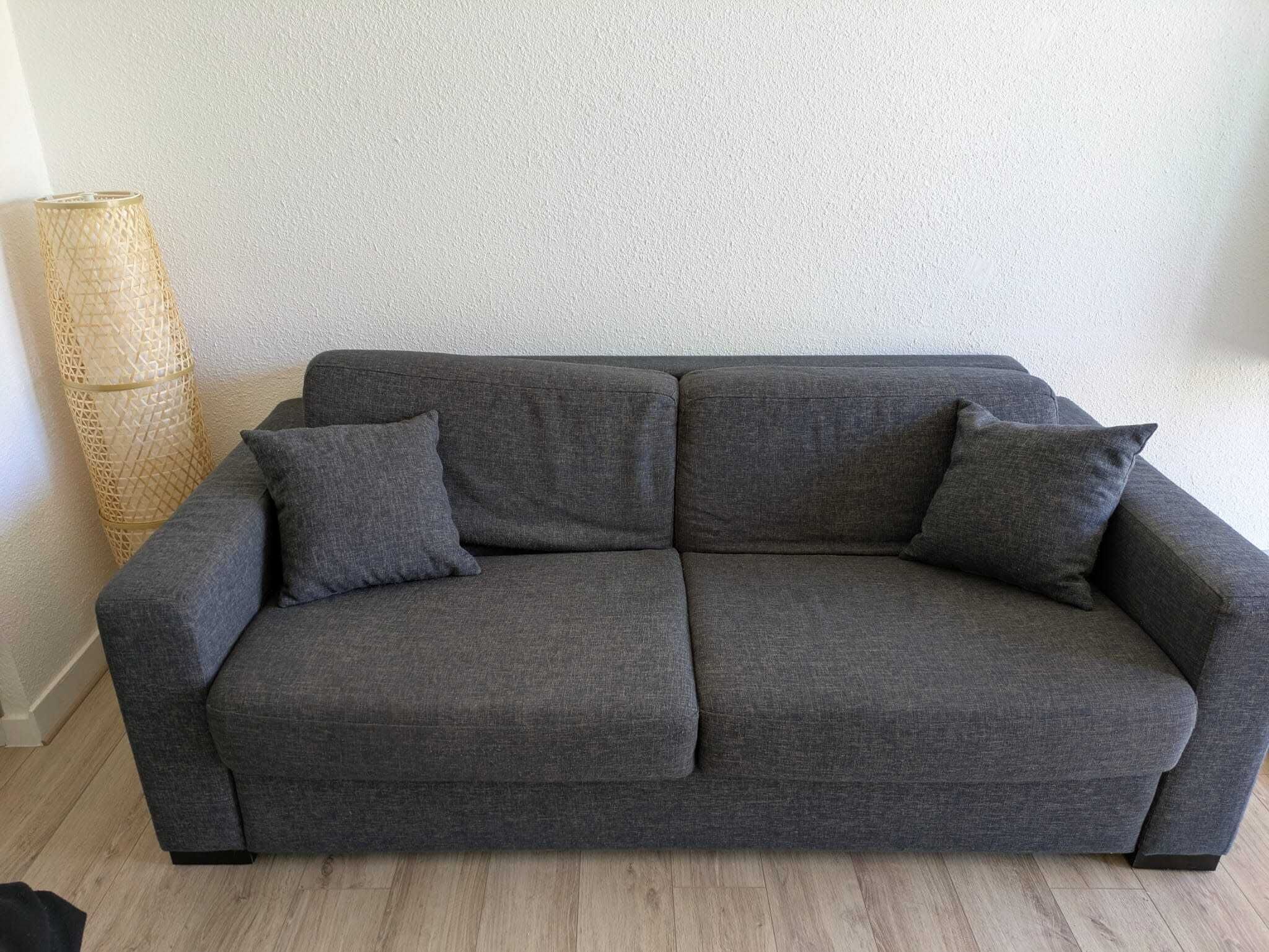 A grey sofa with two cushions in a room with a textured wall and a woven floor lamp.