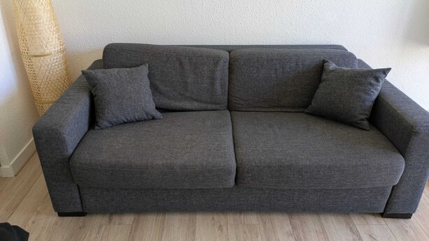 Un sofá gris con dos cojines en una habitación con pared texturizada y lámpara de pie tejida.