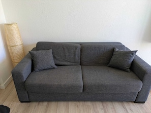 A grey sofa with two cushions in a room with a textured wall and a woven floor lamp. - Studio 4 pers Port Cap d'Agde · Plage (Agde)
