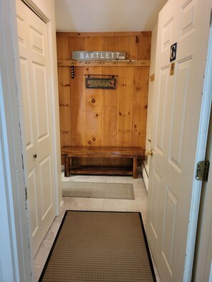 Um corredor com um banco de madeira, uma placa de boas-vindas e uma porta com o símbolo 'G'.