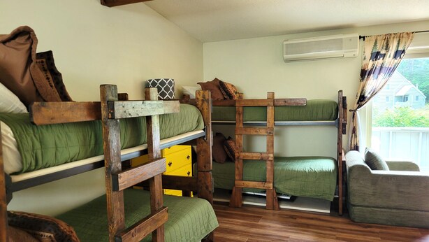 Quarto com beliches de madeira, colchão verde, caixa de armazenamento amarela e janela com cortinas.