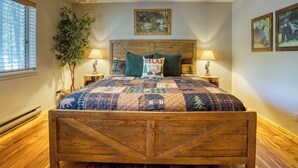 A bedroom with a wooden bed frame, a quilted bedspread, two bedside lamps, a wall-mounted painting, and a potted plant. - Cozy Bear Themed Cabin in Pinetop Country Club (Pinetop Country Club)