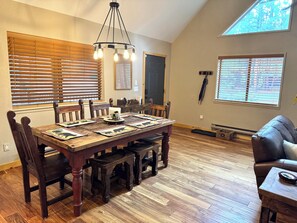 A dining area with a wooden table set for four, flanked by chairs, a chandelier, and large windows with blinds. - Cozy Bear Themed Cabin in Pinetop Country Club (Pinetop Country Club)