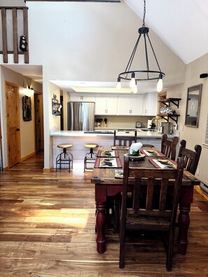 Un coin repas avec une table et des chaises en bois, un luminaire suspendu et une cuisine en arrière-plan.