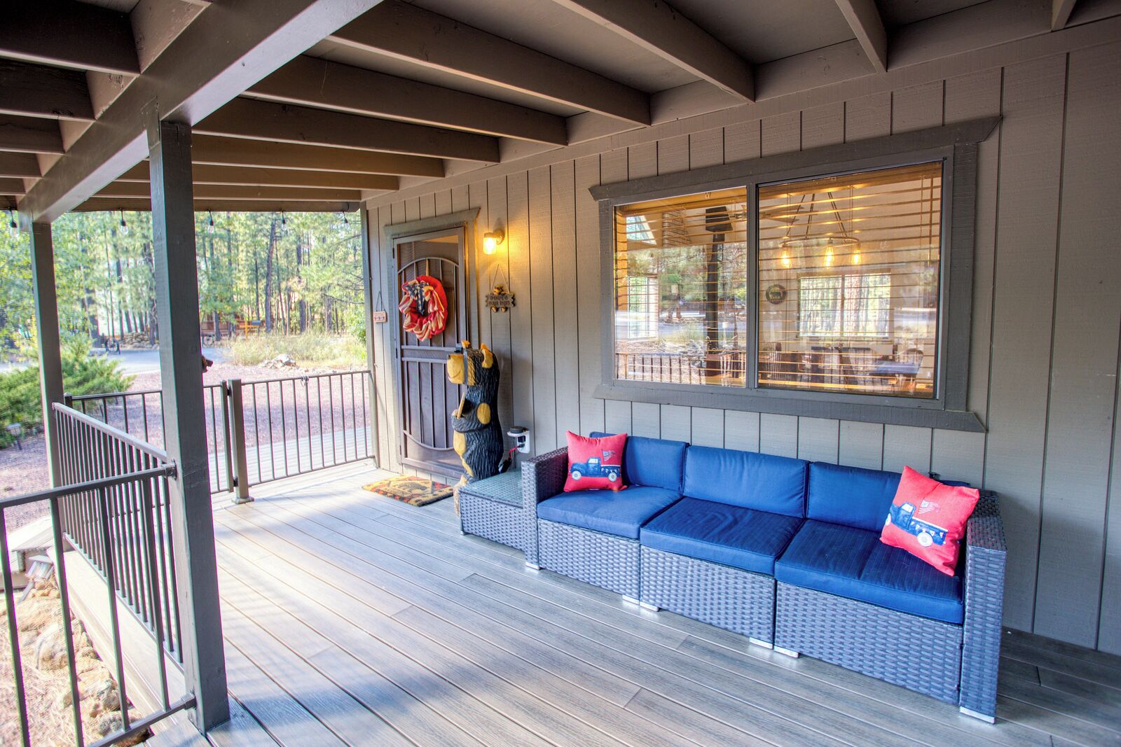 A covered porch with a blue sofa and red pillows, a decorative bear, and a window with blinds.