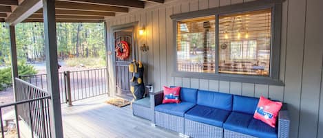 A covered porch with a blue sofa and red pillows, a decorative bear, and a window with blinds.