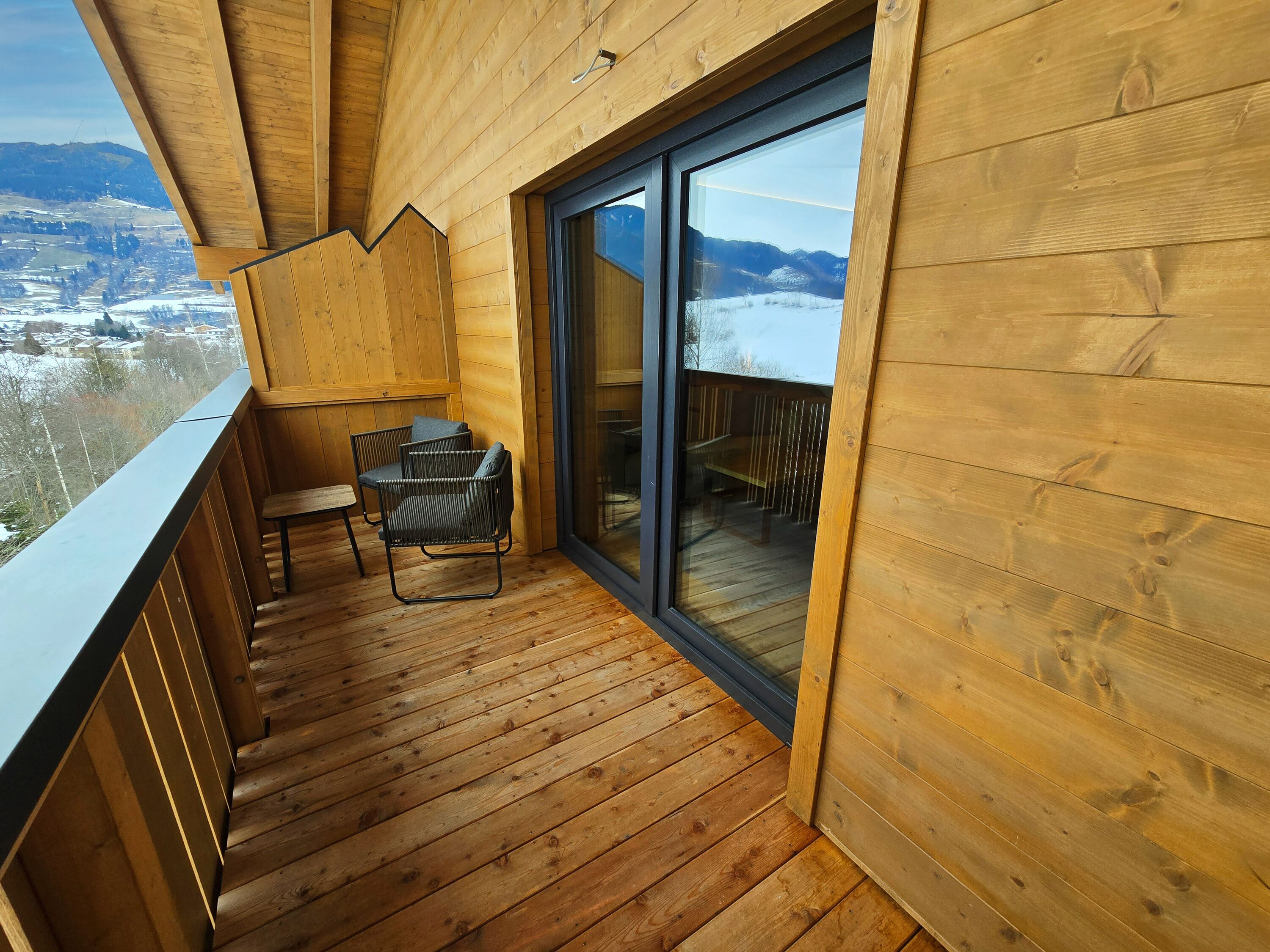 Un balcon en bois avec une porte vitrée donnant sur un espace extérieur, une chaise et une petite table.