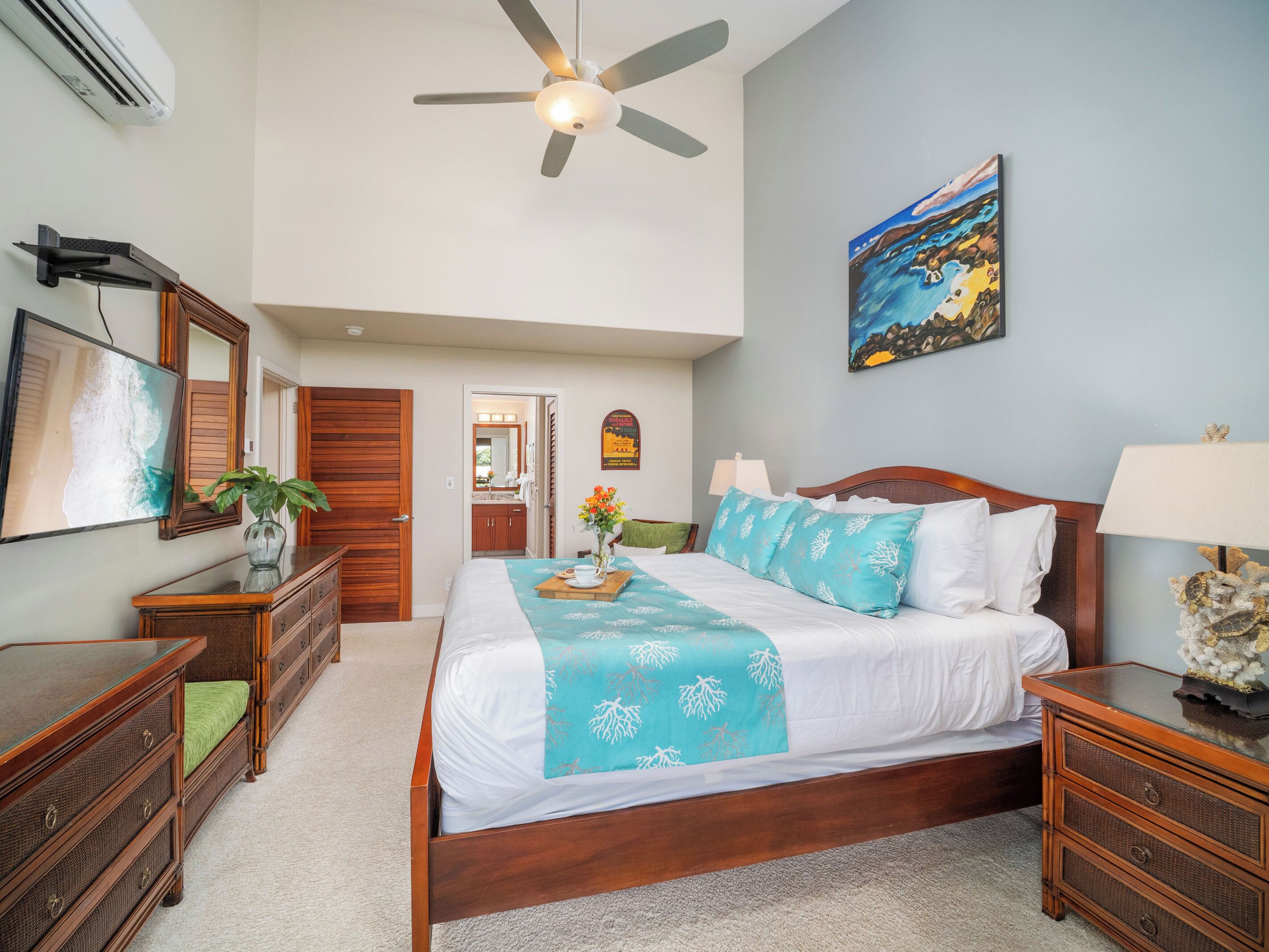 A bedroom with a bed, bedside tables, a ceiling fan, a painting, and a view of the bathroom.