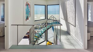 A modern interior with a glass staircase leading to a balcony overlooking a beach. - Iconic Fire Island Pines Oceanfront | Pool, Hot Tub & Sauna (Sayville)