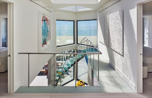A modern interior with a glass staircase leading to a balcony overlooking a beach. - Iconic Fire Island Pines Oceanfront | Pool, Hot Tub & Sauna (Sayville)