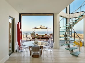 A modern interior with a dining area, clear chairs, a round table, and a view of the ocean. - Iconic Fire Island Pines Oceanfront | Pool, Hot Tub & Sauna (Sayville)