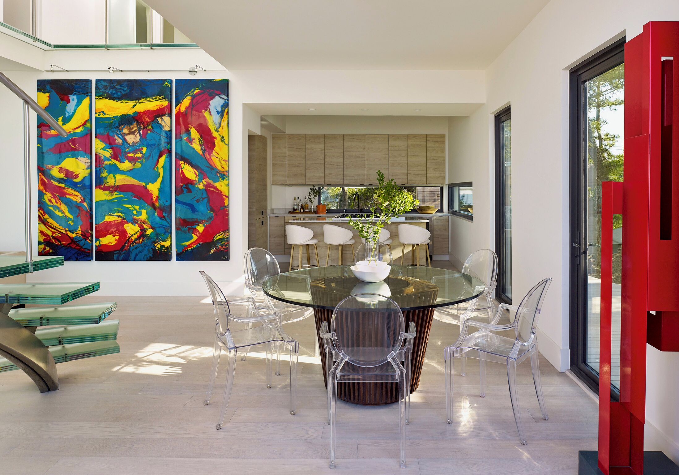 Un espace repas moderne avec une table en verre et des chaises transparentes, une cuisine dotée de meubles en bois et des tableaux abstraits accrochés au mur.