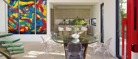 A modern dining area with a glass table and clear chairs, a kitchen with wooden cabinets, and abstract paintings on the wall.