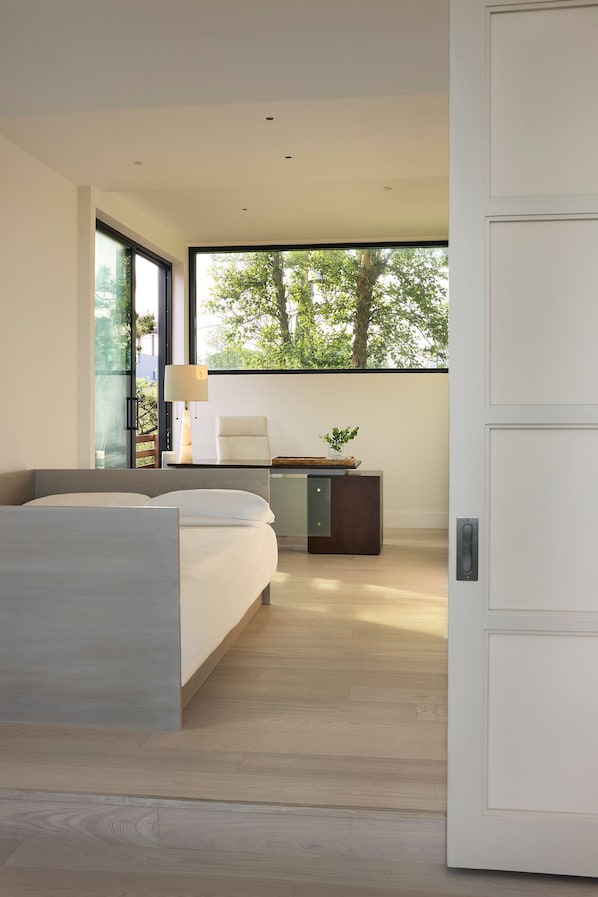 A modern bedroom with a bed, desk, and a view of trees through a large window.