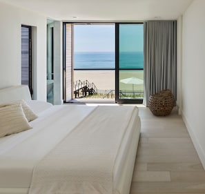 A bedroom with a large bed, a view of the beach, and a balcony with a wicker chair.