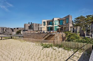 A modern beachfront property with a wooden deck, outdoor seating, and a staircase leading to the entrance.