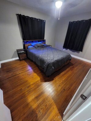 Ein Schlafzimmer mit Bett, Holzboden, Nachttisch und schwarzen Vorhängen.