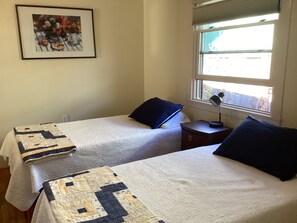 A bedroom with two beds, a nightstand, a lamp, a window with blinds, and a framed picture on the wall. - Stacy’s 2nd st cottages front house (Ashland)