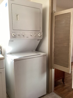 A compact washer and dryer unit with control knobs and a front-loading door, placed in a hallway with a wooden floor and a closed door in the background. - Stacy’s 2nd st cottages front house (Ashland)
