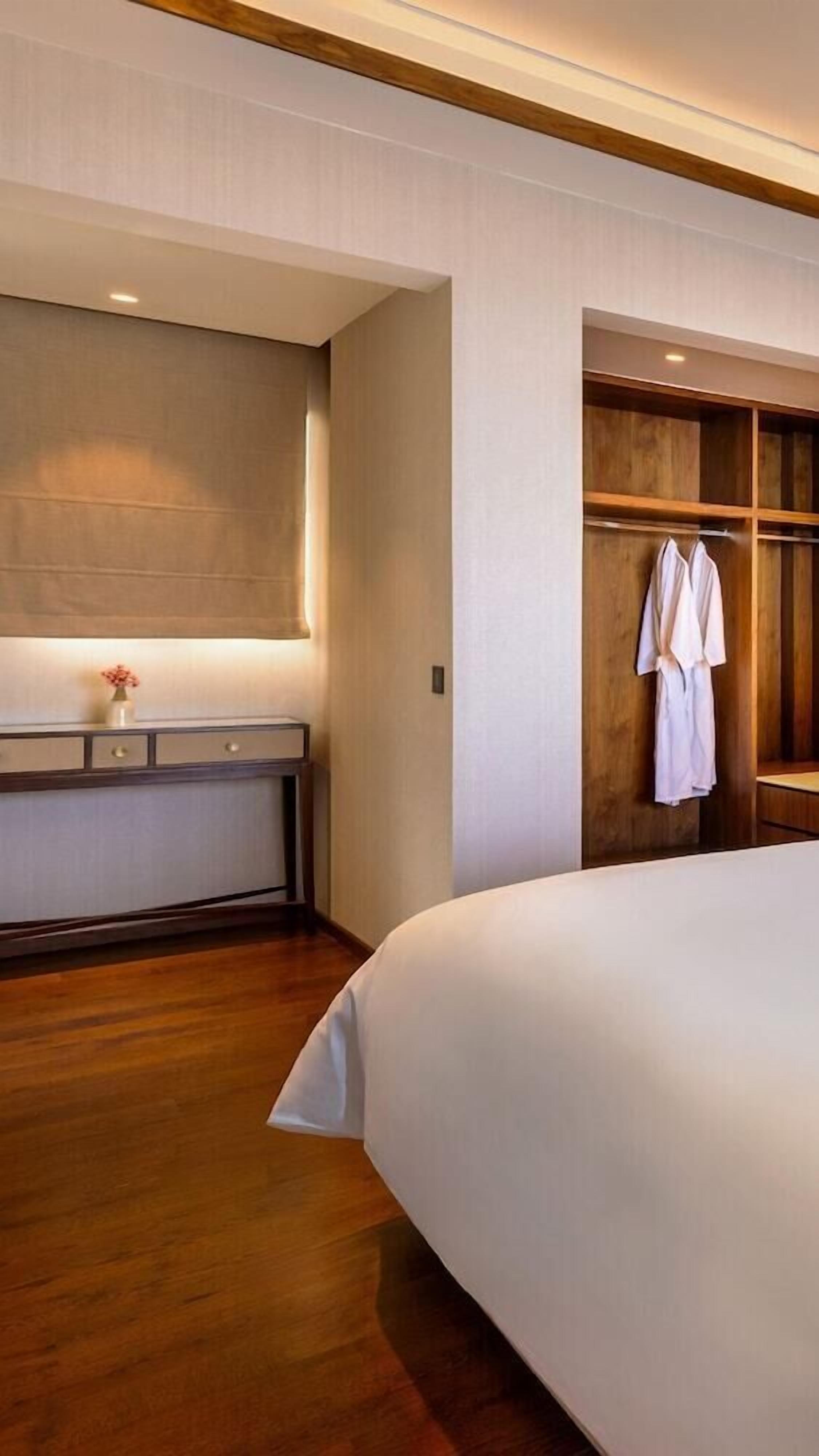 A hotel room with a large bed, a bedside table with a vase, a wooden wardrobe, and a robe hanging on the door.