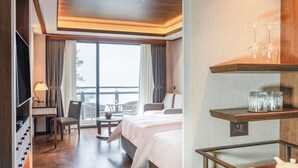 Et hotelværelse med seng, skrivebord, stol, fjernsyn, balkon med udsigt og et hyldesæt med glas og teekande.