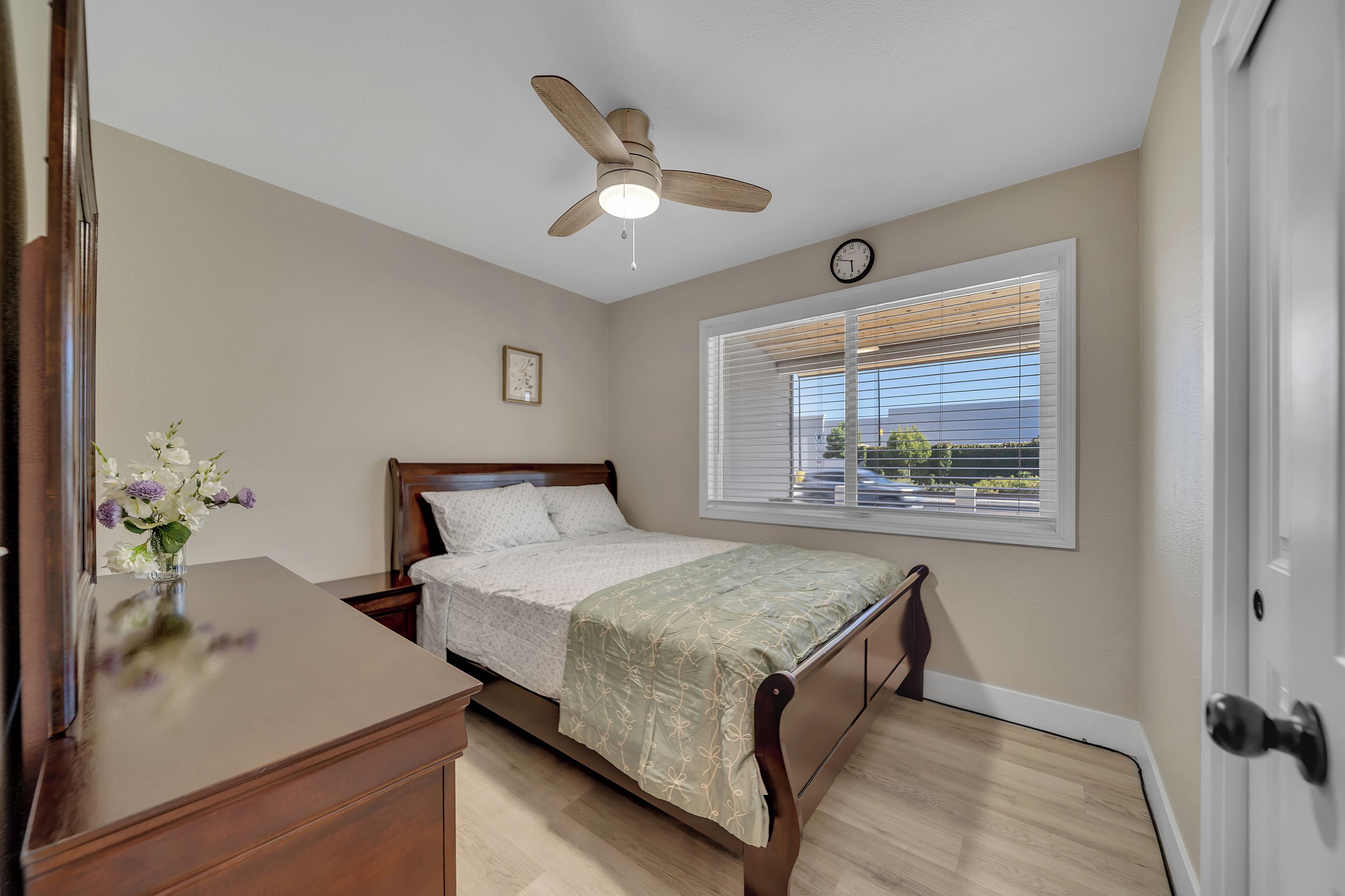 A bedroom with a bed, a ceiling fan, a window with blinds, and a clock on the wall.