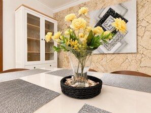Ein Esstisch mit einer klaren Glasvase voller gelber Blumen, einem schwarzen gewebten Korb und einem geometrischen Wandbild.