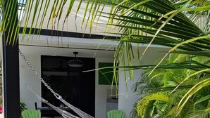 A hammock between two green chairs, a white wall, and a palm tree.