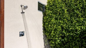 Ein an der Wand befestigter Duschkopf und eine kleine Bedienkonsole neben einer dichten, grünen Hecke.