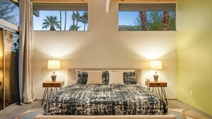 Ein Schlafzimmer mit einem großen Bett, zwei Nachttischen und Blick auf Palmen durch die Fenster.