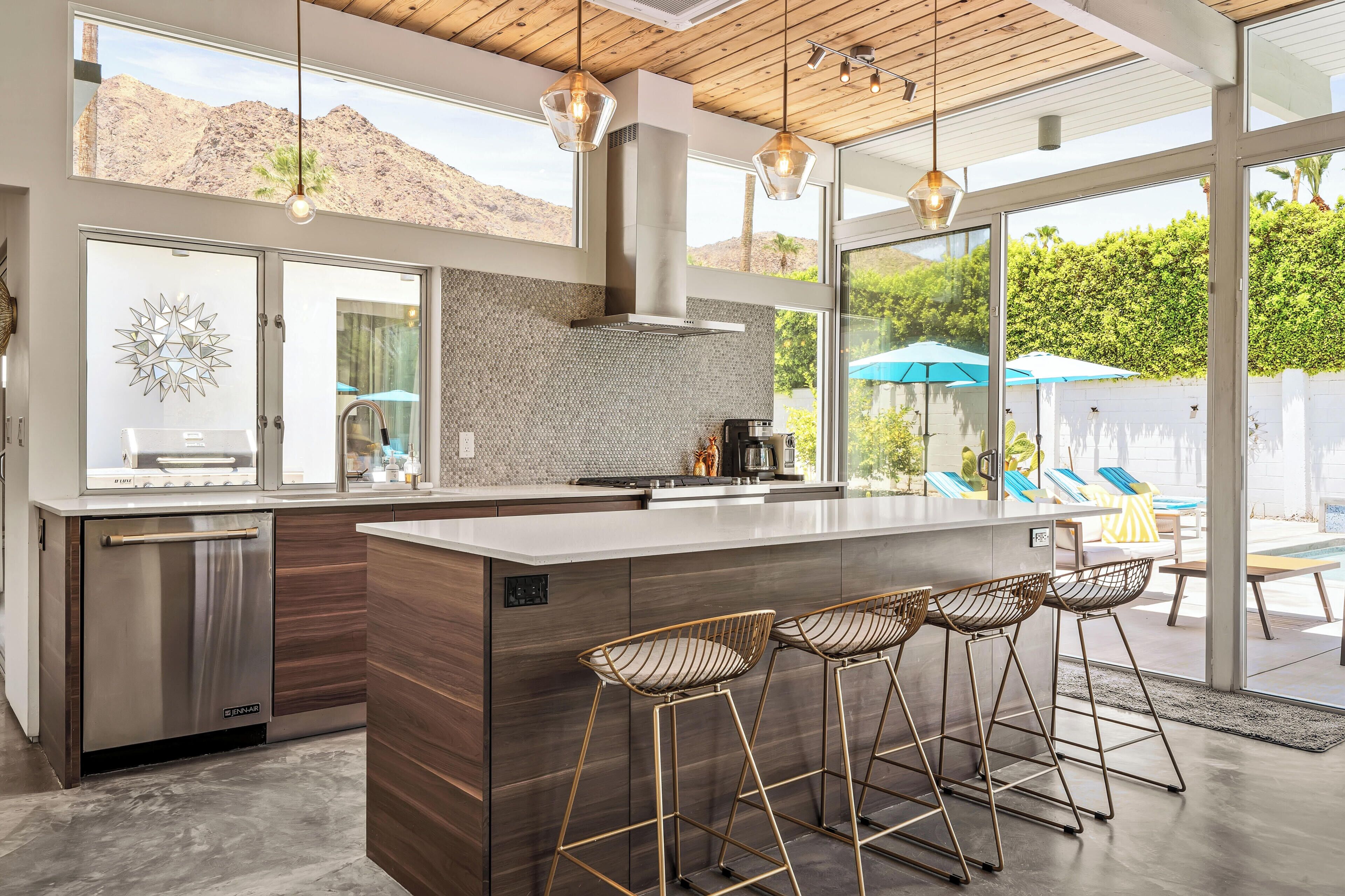 Una cocina moderna con una isla grande, taburetes de bar y vistas a una piscina y follaje.
