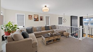 Een woonkamer met een beige bank, een bijzettafeltje en een potplant. Drie ingelijste foto’s hangen aan de muur en er is een plafondlamp.