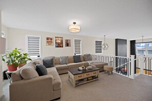 A living room with a beige sofa, a coffee table, and a potted plant. There are three framed pictures on the wall and a ceiling light fixture. - Upscale Show Home | Family House | Workspace (Calgary)