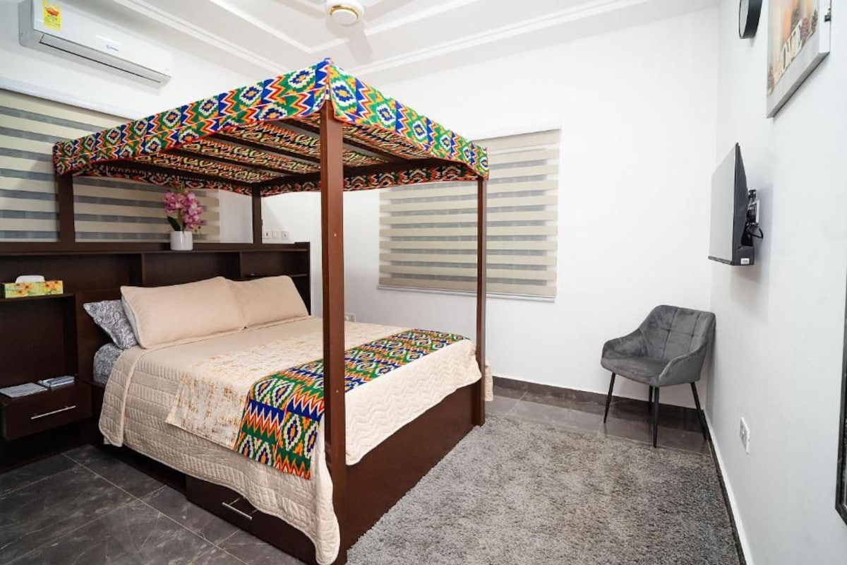 Una cama con dosel y dosel colorido, una mesita de noche con lámpara, una silla y un televisor montado en la pared.