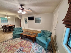 A spacious living room with a wooden coffee table, two patterned armchairs, a dining area with a wooden table and chairs, a ceiling fan, and a large window with a view. - Cozy 2-bedroom house that sleeps 7 (Monroe)
