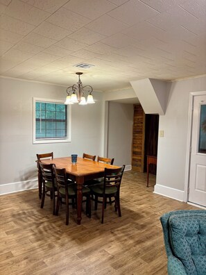 A dining area with a wooden table and chairs, a chandelier, and a window with blinds.