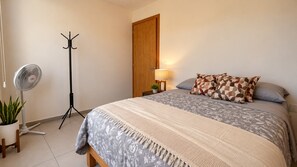 Une chambre à coucher comprenant un lit, un ventilateur sur pied, un porte-manteau, une porte et une table de chevet avec une lampe.
