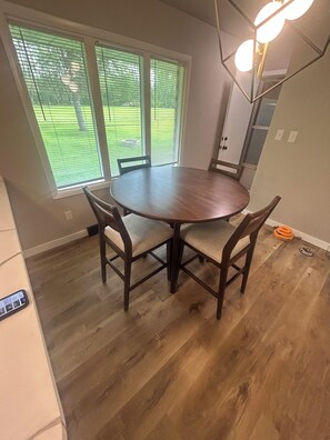 A dining area with a round table and four chairs, large windows with blinds, and a wooden floor.