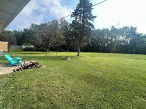 A backyard with a lawn, a blue chair, a fire pit, and a tall tree.