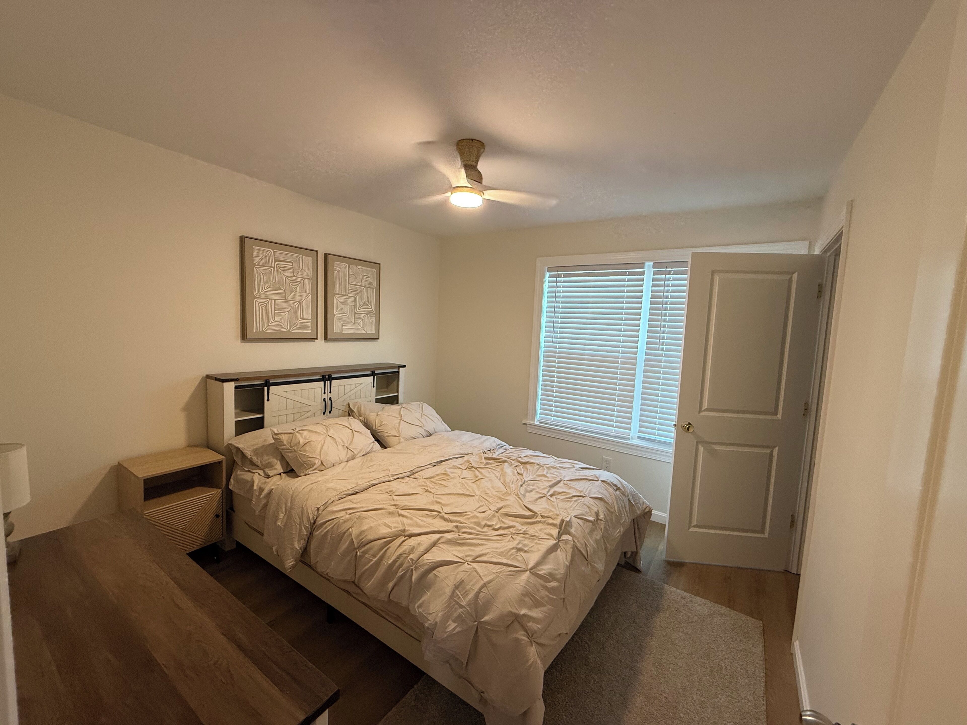 Ein Schlafzimmer mit einem Bett, Nachttischen, einem Deckenventilator, einem Fenster mit Jalousien und zwei Wandbildern.