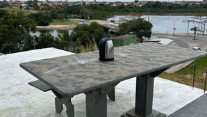 Ein Picknicktisch mit Wasserkocher und Glas, mit Blick auf ein Gewässer und eine Brücke.